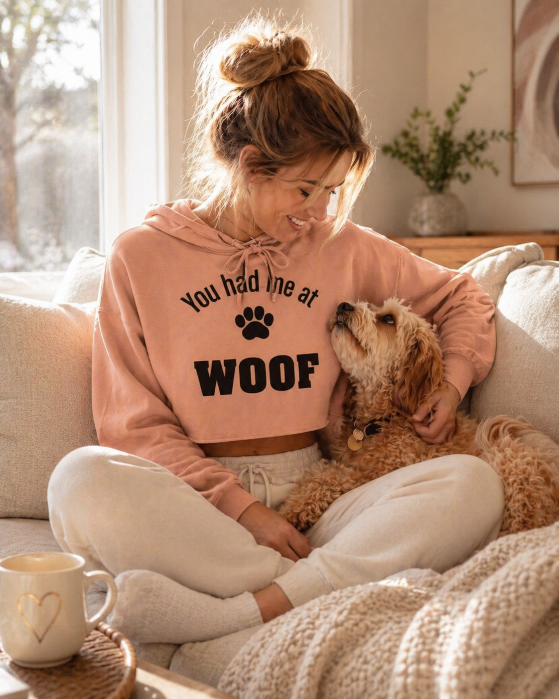 Woman in a pink 'You had me at Woof' hoodie sitting on a couch with a dog.